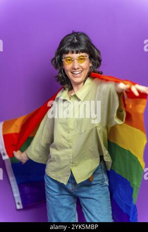 Studio-Porträt mit violettem Hintergrund einer nicht binären lateinischen Frau, die mit lgbt-Regenbogenfahne tanzt Stockfoto