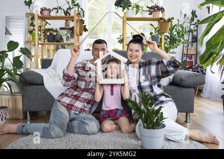 Die traditionelle Familie mit Kind genießt ein neues Zuhause, sitzt auf dem Sofa und hält das Dach. Hypotheken, Versicherung und Schutz, Kauf und Umsiedlung Stockfoto