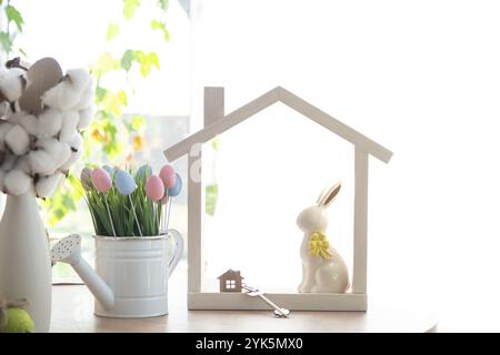 Schlüssel und winziges Haus mit gemütlichem Haus mit Osterdekor mit Kaninchen und Eiern auf dem Tisch der Küche. Bau, Entwurf, Projekt, Umzug in ein neues Haus, Hypothek, Stockfoto