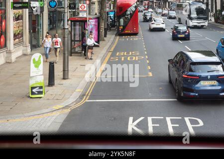 Sie blicken von einem roten Doppeldeckerbus in London hinunter und nähern sich einer markierten Bushaltestelle mit Leuten, die auf der Kensington High Street warten. UK Stockfoto