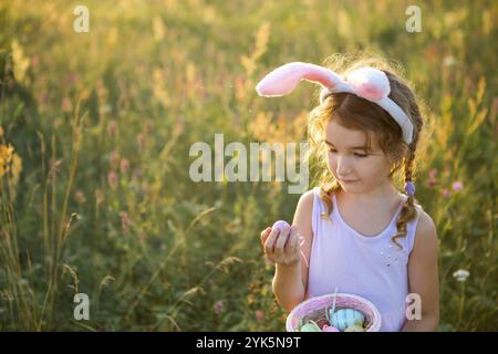 Nettes lustiges Mädchen mit bemalten Ostereiern im Frühling in der Natur auf einem Feld mit goldenem Sonnenlicht und Blumen. Osterferien, Osterhase mit Ohren, Colo Stockfoto