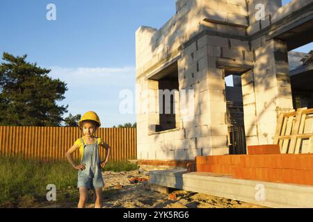 Ein kleines Mädchen in einem gelben Hardhut spielt Baumeister auf der Baustelle ihres zukünftigen Hauses. Träume von Bewegung, die Wahl eines Berufs, Kinder Stockfoto