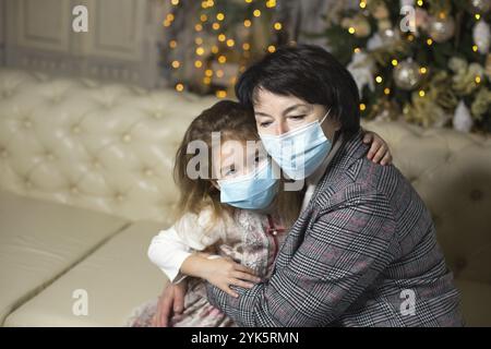 Großmutter und Enkelin auf dem Sofa im Wohnzimmer mit weihnachtlicher Dekoration, die sich in medizinische Masken auf den Gesichtern umarmt. Ein Familienurlaub während der Stockfoto