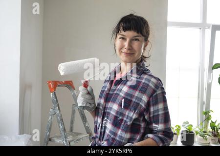 Glückliche Frau mit Malerwalze und weißer Farbe für Wände in Händen Nahporträt. Bauarbeiten und kosmetische Reparaturen im Haus, Wandmalerei, Farbtöne Stockfoto
