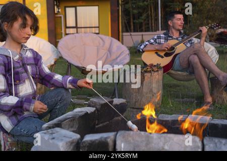 Ein Mädchen in einem karierten Hemd brät Marshmallows auf einem Feuer im Hof des Hauses, Vater spielt Gitarre. Abendliches Familientreffen am Lagerfeuer, Stockfoto