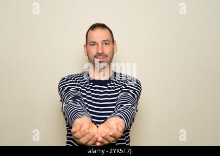 Porträt eines gutaussehenden, freundlichen bärtigen Mannes, der etwas in seinen Händen mit ausgestreckten Armen anbietet. Studio isoliert auf beigefarbenem Hintergrund Stockfoto