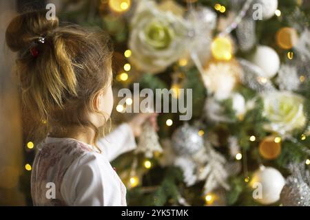 Ein kleines Mädchen greift mit der Hand nach einem Weihnachtsbaumspielzeug. Weihnachtsdekor, warten auf einen Urlaub und ein Wunder. Neujahr. Weiß, rosa und blau verfärben Stockfoto