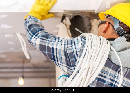 Ein Arbeiter mit Schutzhelm und Handschuhen installiert auf einer Baustelle sorgfältig elektrische Leitungen, während er Schutzausrüstung und ein kariertes Hemd trägt. Stockfoto