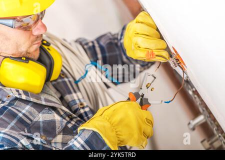 Ein gelernter Elektriker konzentriert sich auf das Anschließen von elektrischen Leitungen, während er Schutzausrüstung trägt und fleißig in einer hellen Wohnumgebung arbeitet Stockfoto