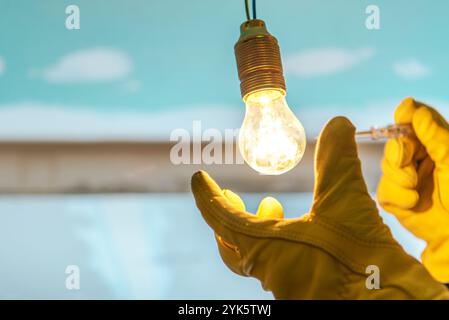 Eine Person, die gelbe Handschuhe trägt, hält eine Glühbirne und passt sie so an, dass helles Licht in einem Arbeitsbereich mit blauem Hintergrund ausstrahlt. Stockfoto