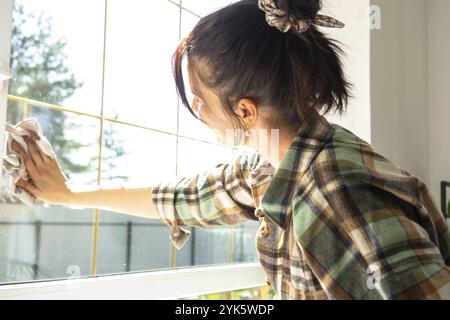 Frau wäscht das Fenster des Hauses manuell mit einem Lappen mit Reinigungsspray und Mopp innen mit weißen Vorhängen. Ordnung und Sauberkeit wiederherstellen Stockfoto