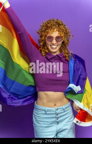 Studio-Porträt mit violettem Hintergrund einer Frau mit lockigem Haar und Sonnenbrille, die lgbt-Regenbogenfahne schwenkt Stockfoto