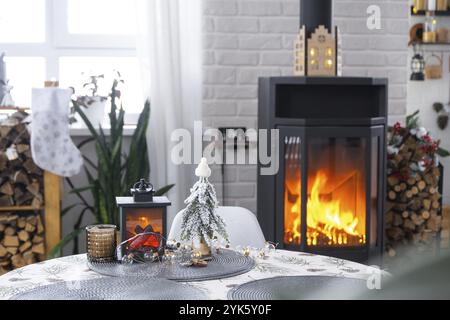 Schwarzer Metallofen, brennender Kamin im weißen festlichen Inneren des Hauses ist für Weihnachten und Neujahr dekoriert, Weihnachtsbaum. Brennholz in t Stockfoto