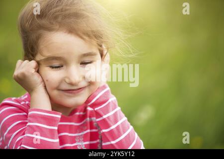 Das kleine Mädchen in einem korallengestreiften T-Shirt auf grünem Hintergrund auf einem Feld hält ihr Gesicht in den Händen und lächelt anstößig. Kindertag, glückliches Kind, env Stockfoto