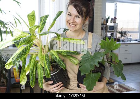 Eine glückliche Frau in einem grünen Haus mit einer Topfpflanze in den Händen lächelt, kümmert sich um eine Blume. Das Innere eines gemütlichen, umweltfreundlichen Hauses, ein Kamin Stockfoto