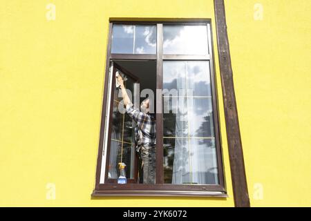 Eine Frau wäscht das Fenster des Hauses manuell mit einem Lappen, einem Sprühreiniger und einem Mopp draußen. Sicherheit in der Höhe, Wiederherstellung von Ordnung und Sauberkeit i Stockfoto