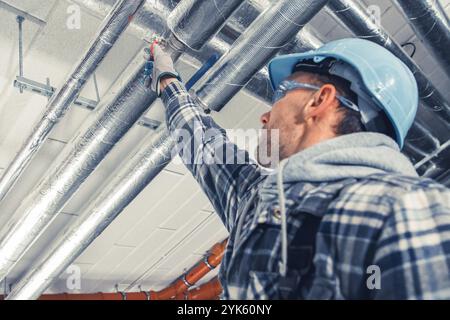 Ein Arbeiter, der Sicherheitsausrüstung trägt, überprüft und stellt die Deckenleitungen in einer Bauumgebung ein, um die ordnungsgemäße Installation und die Einhaltung der Sicherheitsvorschriften zu gewährleisten. Stockfoto