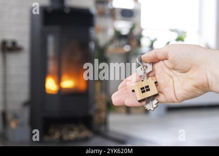 Schlüssel zum Haus mit Schlüsselanhänger in der Hand Besitzer gegen Kamin Herd mit Feuer und Brennholz. Gemütliches Heim. Gebäude, Design, Projekt, Umstieg auf neues Stockfoto