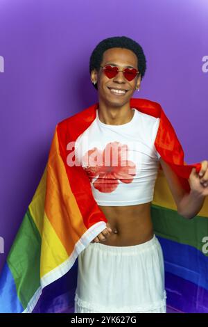 Studio-Porträt mit violettem Hintergrund eines lateinischen schwulen Mannes, eingewickelt in lgtb-Flagge Stockfoto