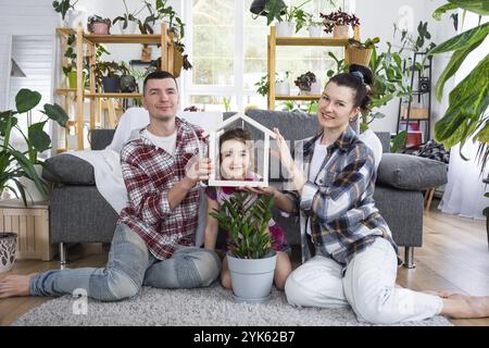 Die traditionelle Familie mit Kind genießt ein neues Zuhause, sitzt auf dem Sofa und hält das Dach. Hypotheken, Versicherung und Schutz, Kauf und Umsiedlung Stockfoto