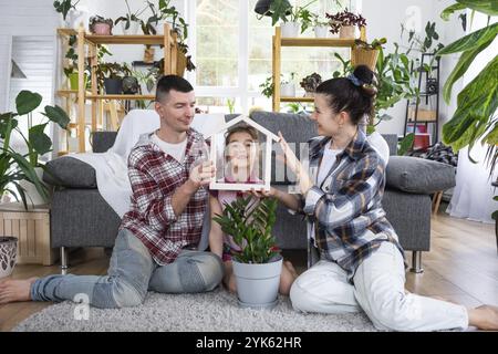 Die traditionelle Familie mit Kind genießt ein neues Zuhause, sitzt auf dem Sofa und hält das Dach. Hypotheken, Versicherung und Schutz, Kauf und Umsiedlung Stockfoto