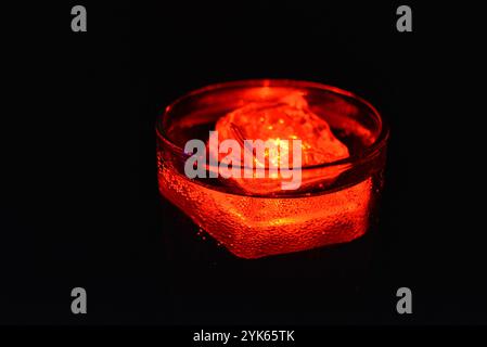 Rot leuchtender Eiswürfel schwimmt in einem Glas Sektglas mit einem Getränk. Plastikgegenstände in Form von Eiswürfeln mit eingebauter Leuchtdiode. Stockfoto