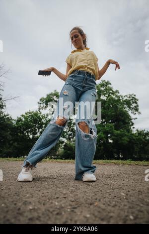 Junge Frau in lässiger Kleidung, die zerrissene Jeans trägt und draußen ein Smartphone hält Stockfoto