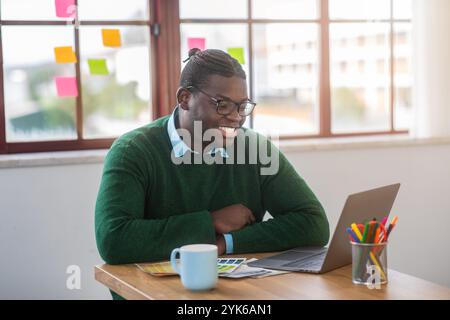 Ein junger Mann sitzt an einem Holztisch und lächelt, während er einen Laptop benutzt Stockfoto