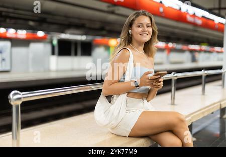 Positives, junges Mädchen, das auf der Plattform sitzt, mit Telefon und kabellosen Kopfhörern auf der Airline Stockfoto