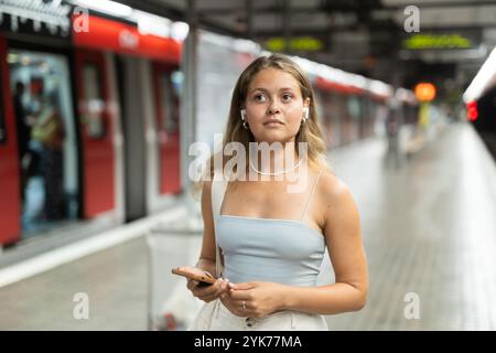 Ein junges Mädchen, das auf der Plattform steht und ein Telefon mit kabellosen Kopfhörern auf Sendung hält Stockfoto