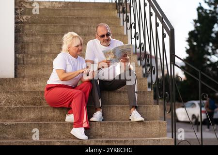 Glückliches Paar beim Erkunden einer Karte, während es im Sommer auf den Stufen der Stadt sitzt Stockfoto