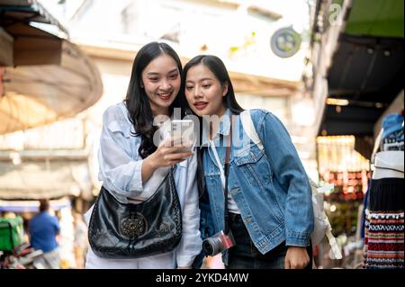 Zwei aufgeregte, fröhliche asiatische Freundinnen genießen es, gemeinsam einen lokalen Markt zu erkunden, einen Ort zu suchen, die Karte auf einem Smartphone zu sehen und ein Fu zu trinken Stockfoto