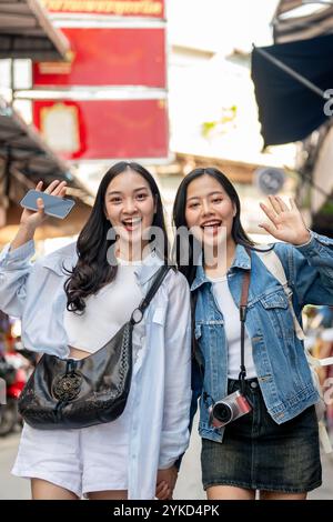 Zwei attraktive, fröhliche asiatische weibliche Touristen in lässiger Kleidung erkunden einen lokalen Markt in Thailand, lächeln und winken der Kamera zu und reisen hinein Stockfoto