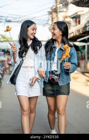 Zwei attraktive, glückliche asiatische Freundinnen, die an einem sonnigen Tag einen Stadtspaziergang genießen, Street Food essen und in Thailand lachen. Urlaub in Thailand Stockfoto