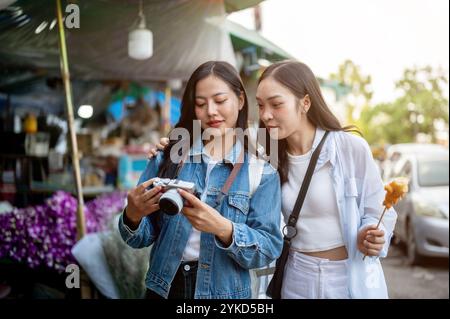 Zwei fröhliche und schöne asiatische Frauen schlendern durch einen Blumenmarkt in Thailand und schauen sich Fotos auf ihrer Kamera an, während sie ihren Urlaub genießen Stockfoto