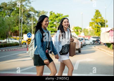 Zwei attraktive, fröhliche junge asiatische Frauen überqueren gemeinsam einen Crosswalk, während sie an einem sonnigen Tag durch eine Stadt in Thailand schlendern und die Stadt erkunden Stockfoto