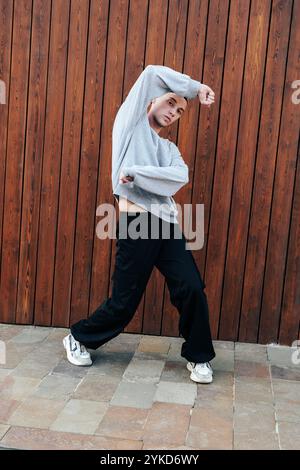 Ein Mann in grauem Pullover und schwarzer Hose tanzt vor einer braunen Holzwand Stockfoto