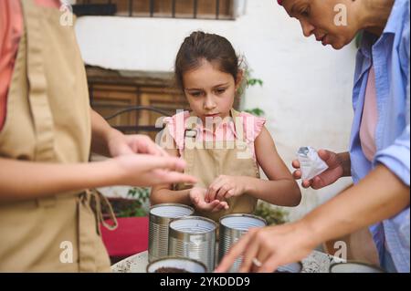 Ein junges Mädchen in einer Schürze wird von einem Erwachsenen geführt, der Samen in Blechdosen pflanzt. Im Freien ist das praktische Lernen mit der Familie und der Natur verbunden. Die sc Stockfoto