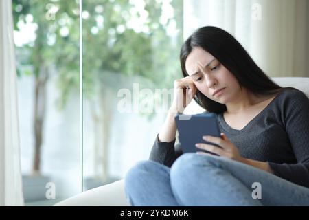 Traurige asiatische Frau, die eBook liest und zu Hause auf einer Couch sitzt Stockfoto