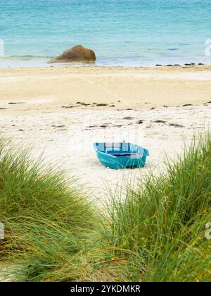 Ein kleines blaues Boot ruht an einem Sandstrand von Pontusval mit Gras im Vordergrund und dem Meer im Hintergrund. Stockfoto