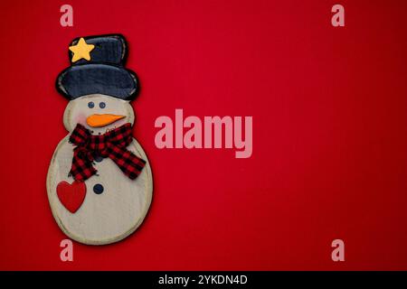 Ein hölzerner Schneemann mit schwarzem Hut, rotem Schal und Herz, vor einem leuchtend roten Hintergrund, der festliche Stimmung verleiht. Stockfoto