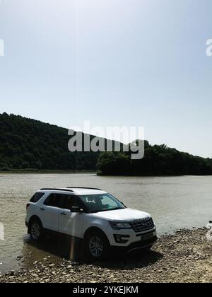 Kiew, Ukraine - 04. April 2021: Ein weißer Ford Explorer SUV steht an einem felsigen Fluss unter klarem Himmel, umgeben von üppig grünen Wäldern, Kreat Stockfoto