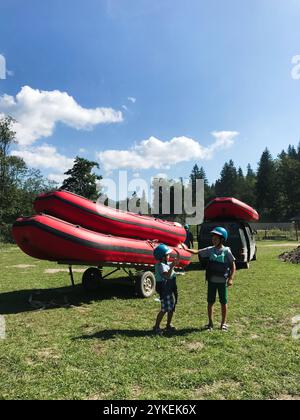 Zwei Kinder mit Helmen und Schwimmwesten stehen neben roten Flößen und einem Truck bereit für ein Rafting-Abenteuer. Heller, sonniger Tag unterstreicht die Spannung Stockfoto