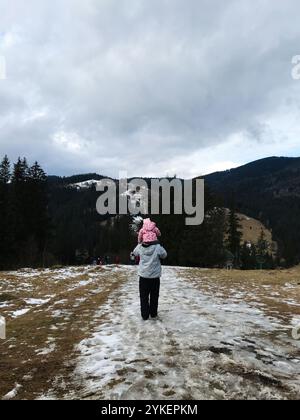 Ein Elternteil trägt ein Kind auf den Schultern während einer Winterwanderung durch eine malerische Bergwelt. Schneebedeckte Gipfel und bewaldete Landschaft schaffen eine Stockfoto