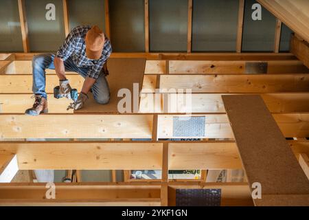 Ein Zimmermann installiert sorgfältig Bodenbeläge in einem neuen Haus, indem er die Bretter mit Elektrowerkzeugen fixiert, während sie von Holzbalken und Sonnenlicht umgeben sind. Stockfoto