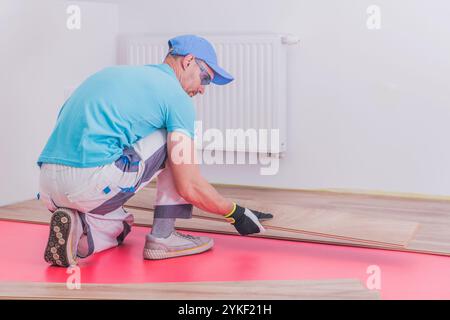 Ein Mann legt sorgfältig Holzdielen auf eine hellrote Unterlage und konzentriert sich dabei auf die präzise Installation in einem neu renovierten Raum. Stockfoto