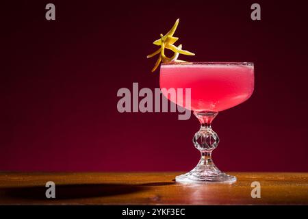 Kosmopolitischer Cocktail, präsentiert in einem Glas mit orangefarbener Verzierung vor einem satten roten Hintergrund, fängt dieses Bild die Essenz der Sophisti ein Stockfoto