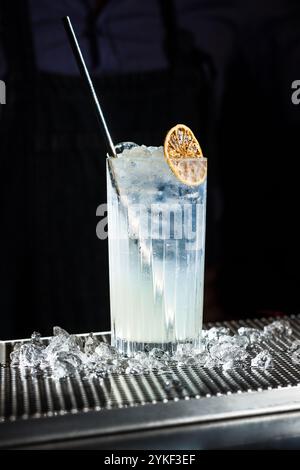 Ein erfrischender Tom Collins Cocktail, serviert in einem hohen Glas mit Eiswürfeln und garniert mit einer getrockneten Zitrusscheibe, die perfekt auf einer Bartheke präsentiert wird Stockfoto