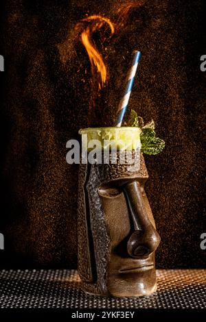 Ein markanter Tiki-Cocktail ist mit einer feurigen Zierde verziert, serviert in einem aufwendig gestalteten Becher mit Limette und Minze, der eine optisch fesselnde und faszinierende Note bietet Stockfoto