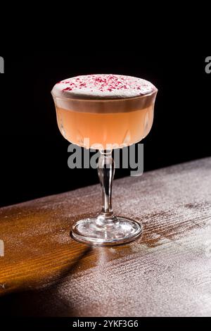 Ein wunderschön gefertigter Pisco Sour Cocktail, gekrönt mit schaumigem Schaum und einem Hauch von rosa Garnituren, sitzt elegant auf einem rustikalen Holztisch vor einem d Stockfoto